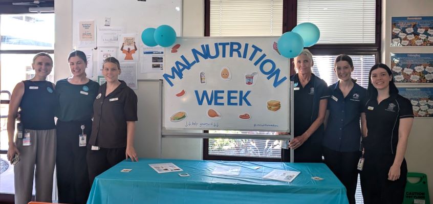 Healthcare staff standing next to display board with signage for Malnutrition Week