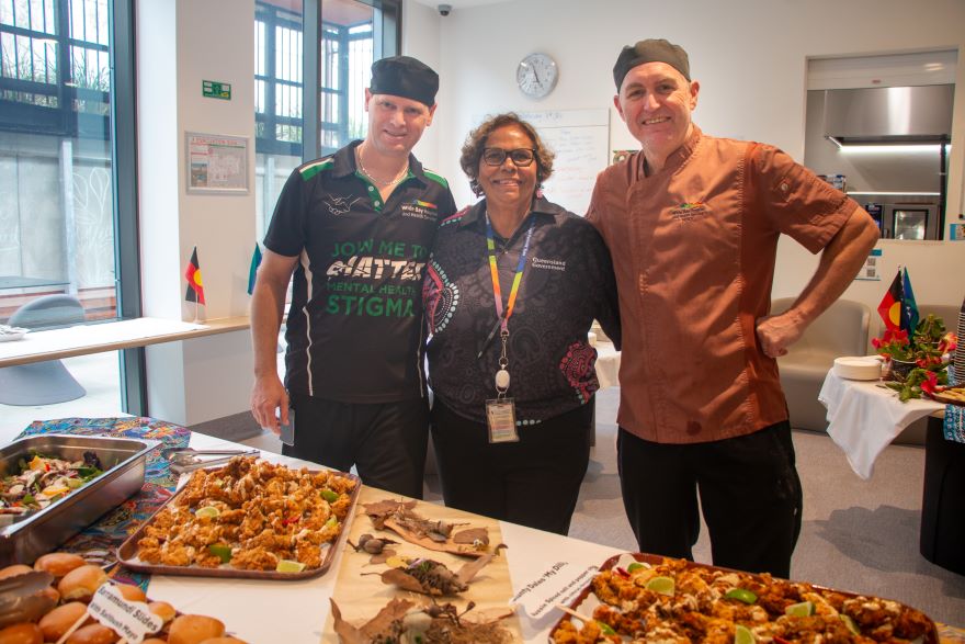 Three hospital staff members standing next to selection of food prepared for NAIDOC week.