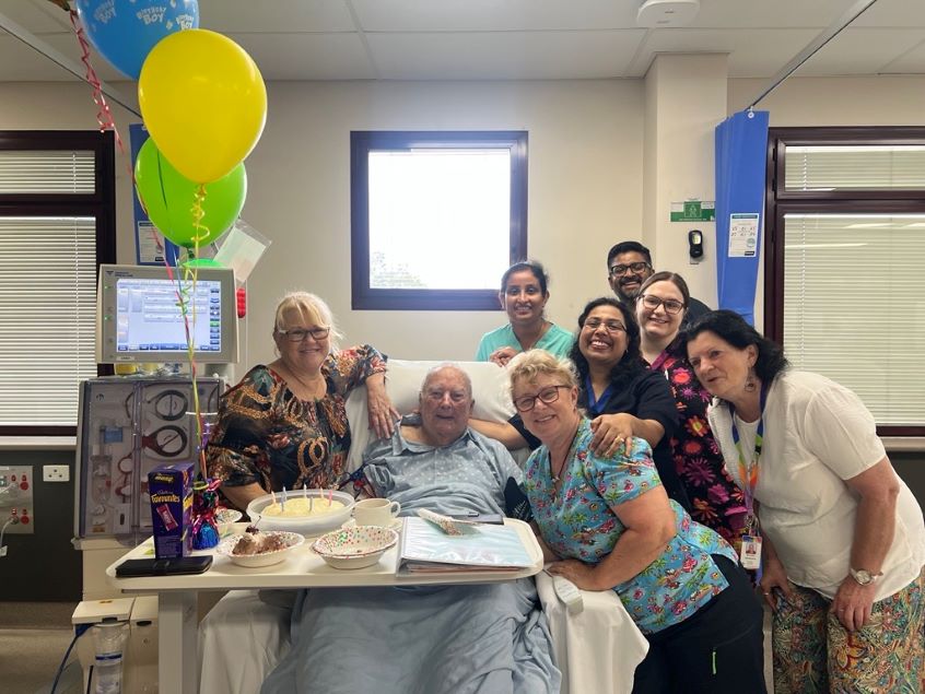 Male patient in hospital bed with balloons and surrounded by nursing staff